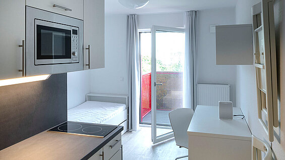 Durchblick von der Küche ins Zimmer; die Küche ist schlicht in weiß und grau, die Tür zum kleinen Balkon ist offen