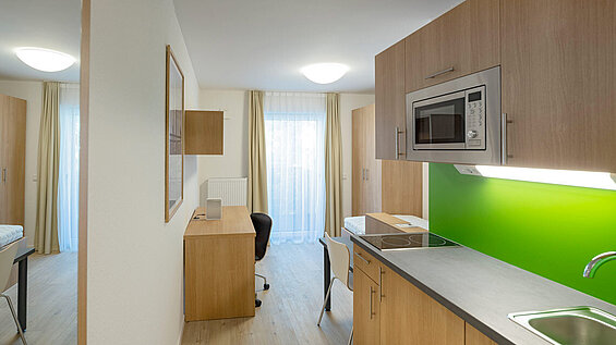 Durchblick von der Küche ins Zimmer; die Möbel sind aus hellem Holz, die schmale Küchenzeile hat eine grüne Rückplatte